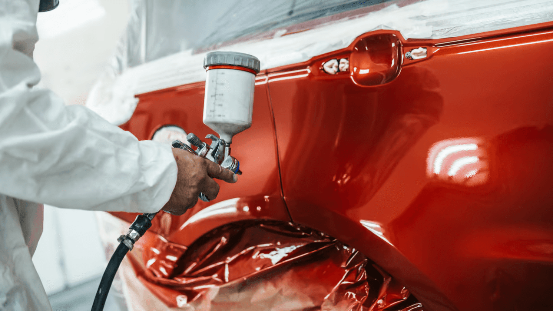 Close-up of a person in protective gear spray painting a red car part in a workshop.